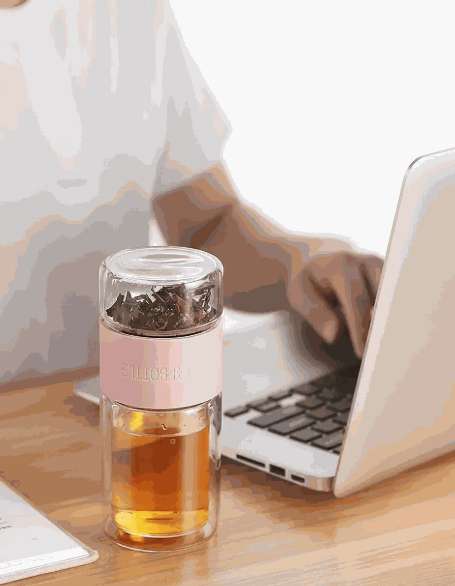 Clear glass tea infuser with pink lid on a wooden desk next to a laptop.