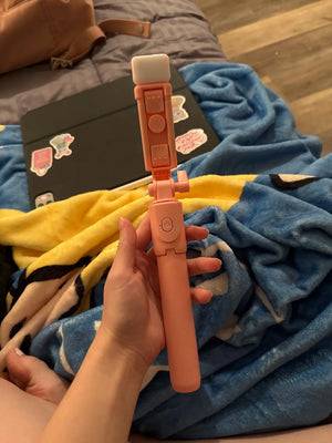 Hand holding an orange selfie stick with a blurred background of a bed with colorful blankets.