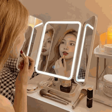 Person applying lipstick in front of a illuminated vanity mirror with various makeup items on a table.