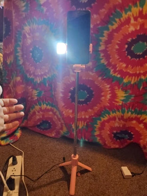 Pink selfie stick with smartphone holder on a carpeted floor against a tie-dye curtain background.
